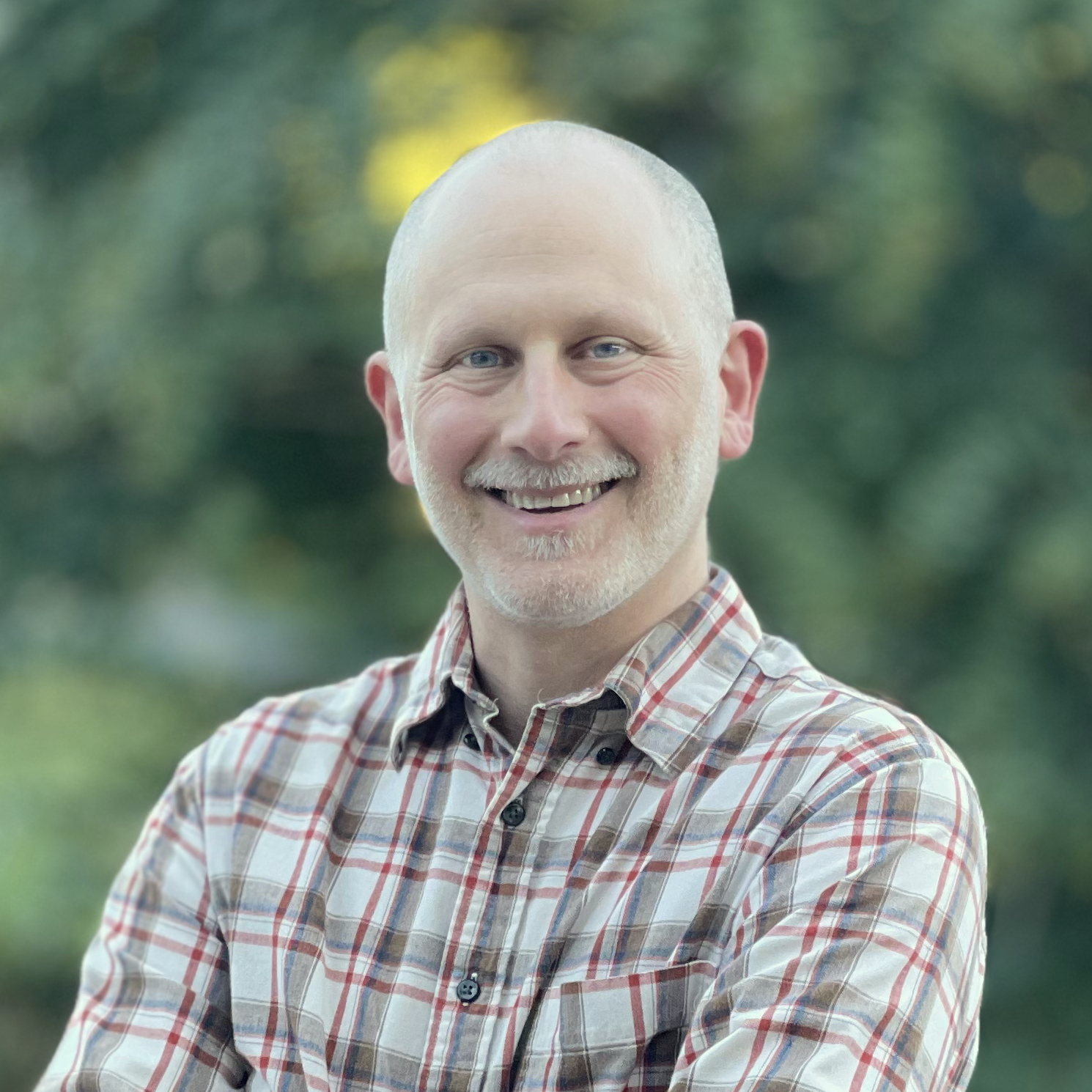 An image of a man with a shaved head, a slight mustache who is wearing a plaid shirt smiling in front of greenery